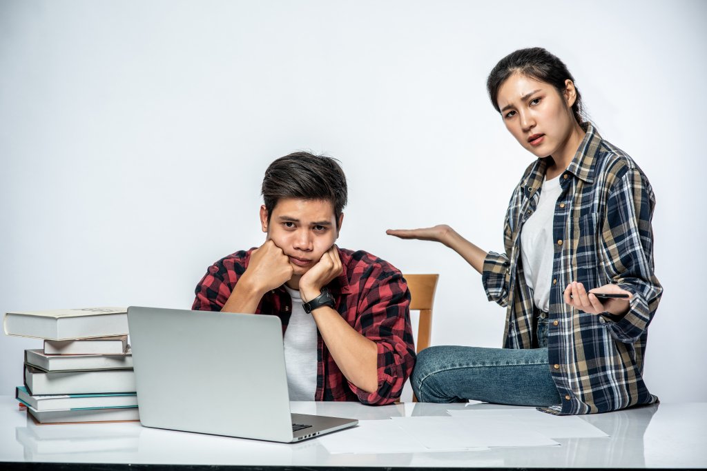 "Female student guiding a frustrated male student with laptop and study materials on the table."
